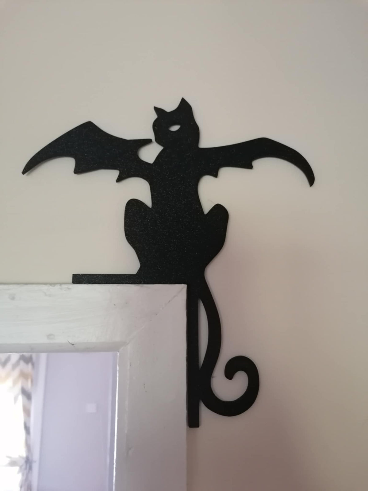 Black silhouette of a cat with bat wings perched on a door frame against a light background.