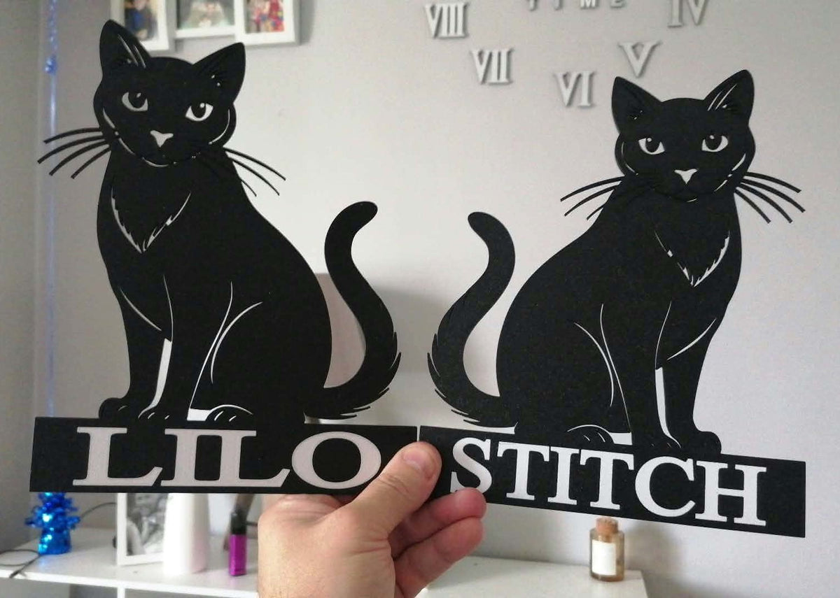 Personalised black cat silhouette wall art featuring custom pet names "Lilo" and "Stitch" beneath detailed cat designs. The black 3D printed cat plaques display expressive facial features and elegant curved tails, shown being held indoors against a light-coloured wall with decorative Roman numeral wall clock elements in the background.