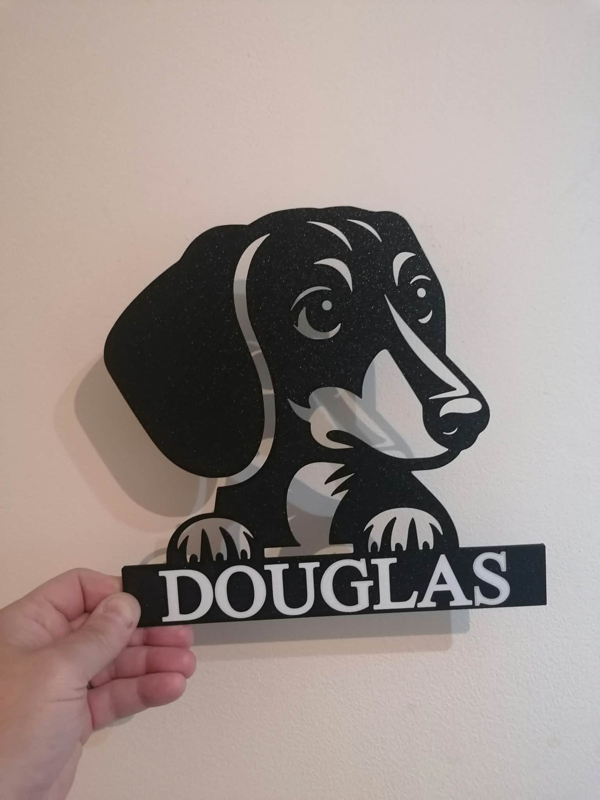Personalised dachshund wall art featuring a black sausage dog silhouette with floppy ears, expressive eyes and front paws resting above the custom name "DOUGLAS". The 3D printed design is shown being held against a light-coloured wall, demonstrating the optional personalised name feature available with the artwork.