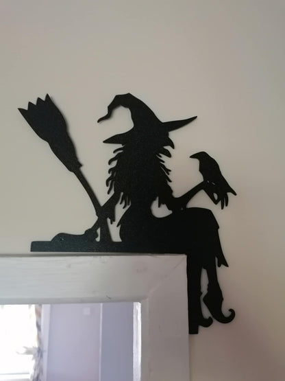 Black witch silhouette corner wall art mounted on the top corner of a white door frame. The 3D printed design features a seated witch wearing a pointed hat, holding a broomstick, with a raven perched beside the figure. The gothic silhouette extends over both sides of the corner, creating a striking Halloween and witchcore décor accent against a light-coloured wall.