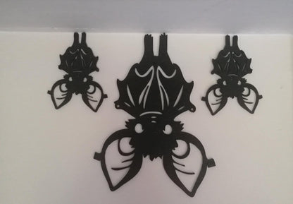 Black silhouette trio of stylised bats hanging upside down, featuring bold wings and decorative detailing on a white background.